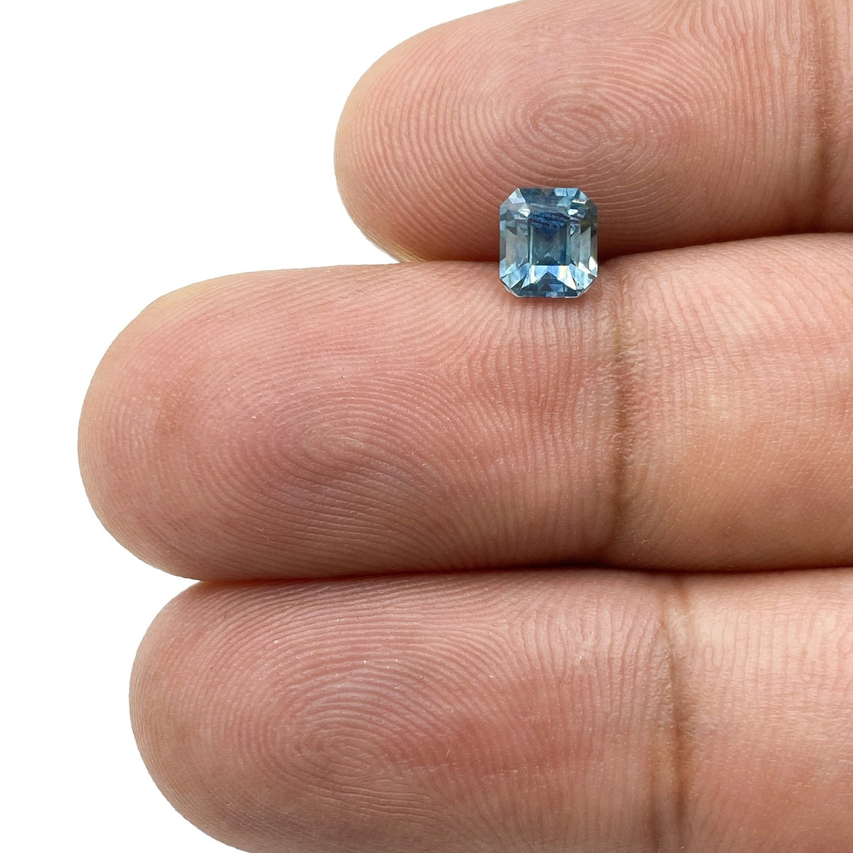 0.94ct Asscher cut blue Montana sapphire held between fingers, showcasing its square shape and faceted blue gemstone surface.