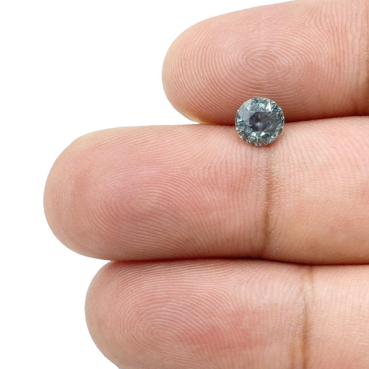 1.01ct round brilliant cut blue Montana sapphire held between fingers, showing facets and grey-blue tones.