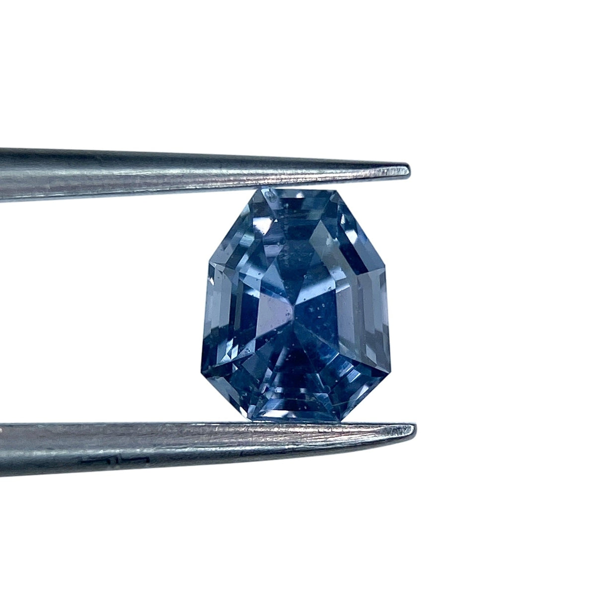 1.01ct shield-shaped blue sapphire with a step cut, held in metal tweezers against a white background.