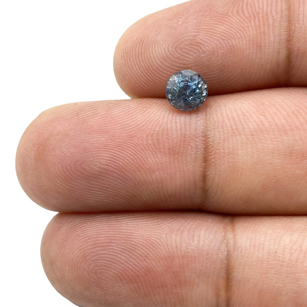 1.02ct round brilliant cut blue Montana sapphire held between fingers, showing deep blue tones and faceted sparkle.