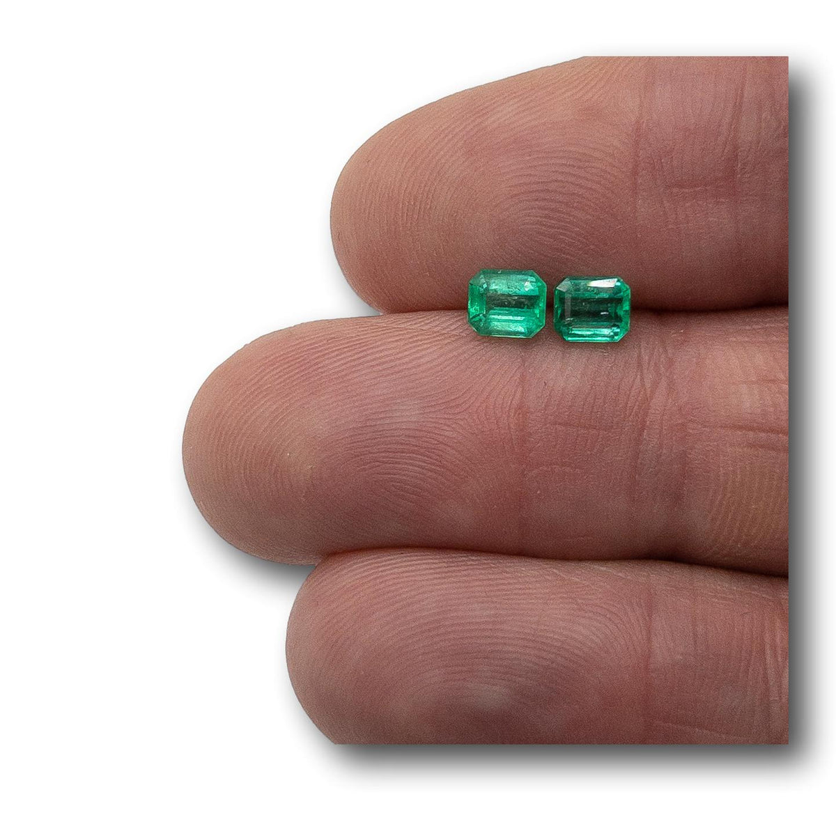 Pair of 1.05ct Muzo emeralds with step-cut emerald shapes held on fingers.