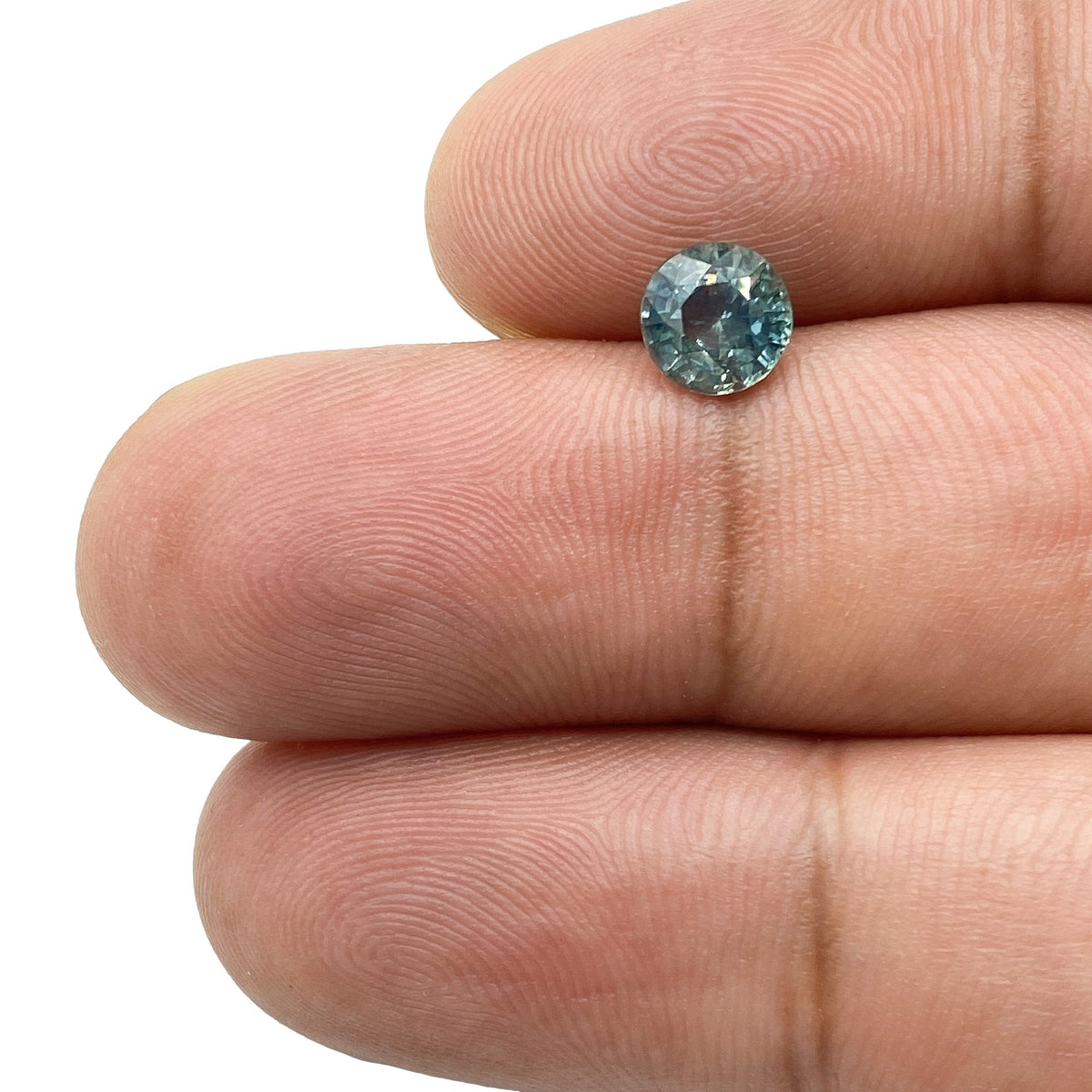 1.06ct round brilliant cut blue Montana sapphire held between fingers, showcasing its faceted sparkle and teal-blue hue.