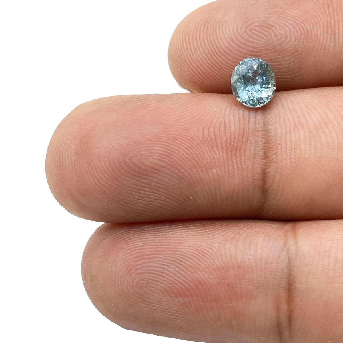 1.07ct brilliant cut oval blue Montana sapphire held between two fingers, showing its sparkling facets and light blue color.