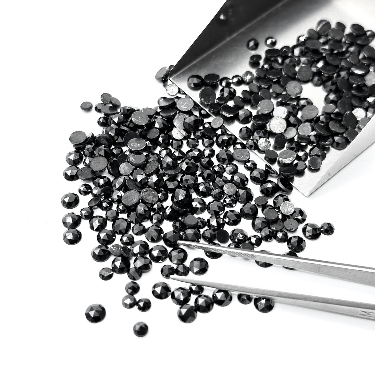 Small, round black diamond melee with a rose cut finish, poured from a metal scoop with tweezers on a white background.