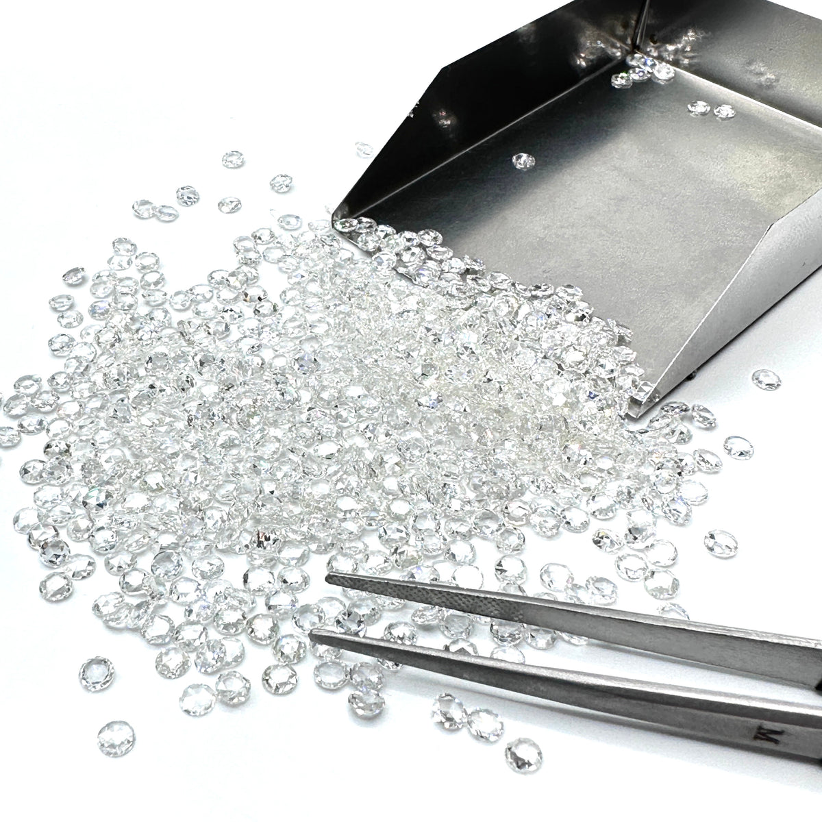 Small, sparkling white rose cut diamond melee stones scattered with a metal jewelry scoop and tweezers on a white surface.